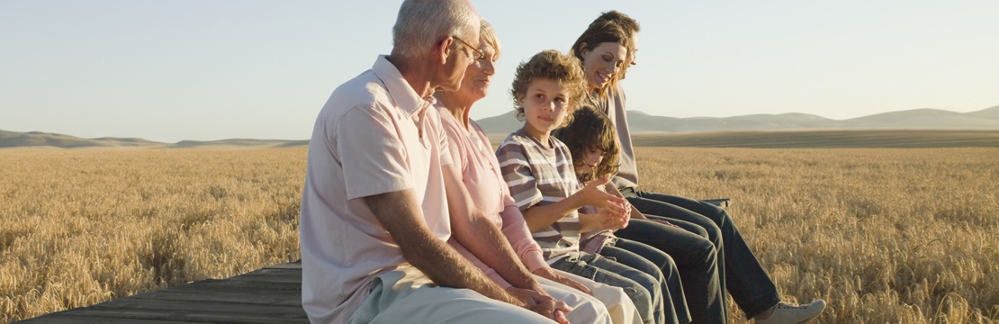 Multigenerational family sitting on a wooden platform at the edge of a golden wheat field, with mountains visible in the distance under a clear sky.